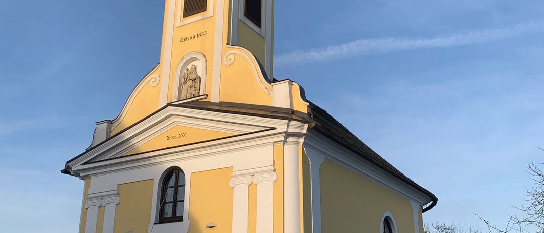 Eine kleine gelbe Kirche mit einem Turm und einer Uhr auf dem Glockenturm, erbaut im Jahr 1833. Sie hat eine Statue an der Vorderseite und zwei Bänke davor.