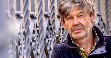 Ein Mann mit grauen Haaren, der eine blaue Jacke trägt und eine Sonnenbrille auf dem Kopf hat, steht vor einem Metallzaun.