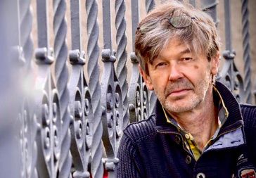 Ein Mann mit grauen Haaren, der eine blaue Jacke trägt und eine Sonnenbrille auf dem Kopf hat, steht vor einem Metallzaun.