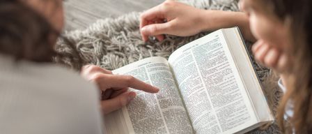 Zwei Personen lesen gemeinsam ein Buch, eine zeigt auf eine Seite, während die andere zusieht. Das Buch ist geöffnet und zeigt Text und ein Bild. Sie sitzen auf einem Teppichboden.