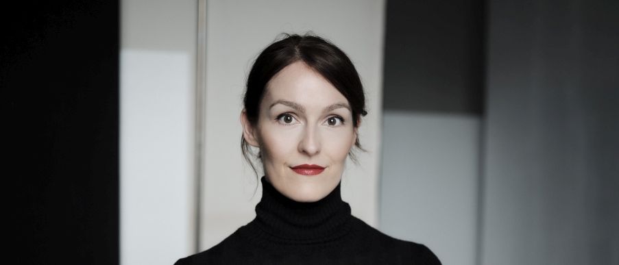 Eine Frau steht in einem Raum mit einer weißen Wand hinter ihr, trägt ein schwarzes Rollkragenshirt und lächelt. Sie hat dunkles Haar und roten Lippenstift.