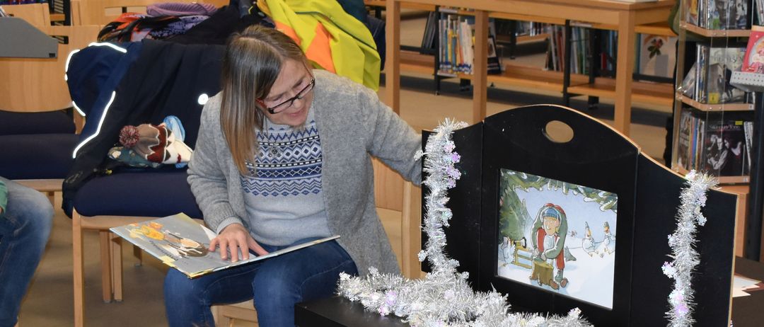 Eine Frau liest Kindern vor, die in einem Kreis um einen geschmückten Tisch mit einem Fernsehbildschirm sitzen. Im Hintergrund sind mit Büchern gefüllte Regale zu sehen.