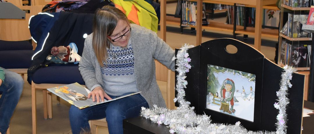 Eine Frau liest Kindern in einer Bibliothek ein Buch vor. Sie sitzen um einen Tisch mit Bildschirm und Tinsel. Dahinter sind Regale mit Büchern gefüllt.