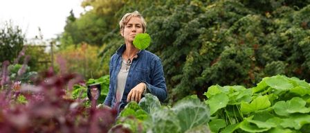 Eine Frau steht in einem Garten mit grünen Blättern im Mund und hält eine Schaufel in der linken Hand.