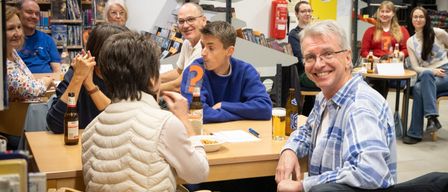 Mehrere Personen sitzen in einer Bibliothek um einen Tisch herum, lächeln und unterhalten sich. Ein Mann in einem blauen Pullover sieht nachdenklich aus, während andere Getränke und Essen halten.