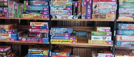 Ein Holzregal, gefüllt mit verschiedenen Brettspielen, darunter Titel wie Zug um Zug, Carcassonne und andere, ordentlich in Reihen angeordnet.