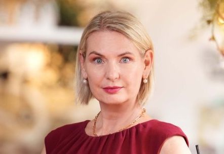 Eine Frau mit kurzen blonden Haaren, blauen Augen und einem warmen Lächeln. Sie trägt ein weinrotes Oberteil, eine goldene Halskette und Ohrringe. Sie scheint sich in einem Raum mit einer weißen Wand und einem dekorativen Element über ihrem Kopf zu befinden.