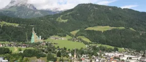 Luftaufnahme einer kleinen Stadt in den Alpen mit einem grünen Feld, Bergen und einem großen Gebäude.