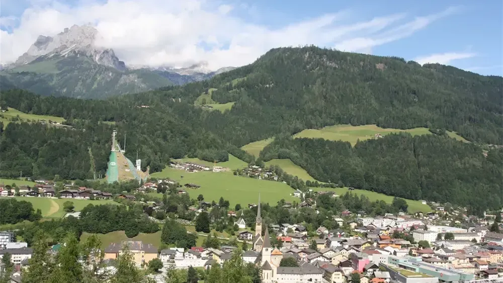 Luftaufnahme einer kleinen Stadt in den Alpen mit einem grünen Feld, Bergen und einem großen Gebäude.