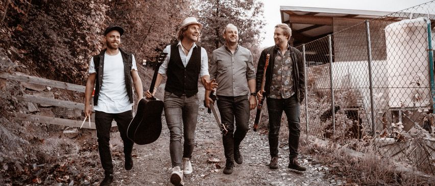 Vier Männer, jeder mit einer Gitarre, gehen gemeinsam auf einem Kiesweg. Sie tragen lässige Kleidung, einer trägt einen Hut und ein anderer eine Halskette. Dahinter ist ein Gebäude mit einem Zaun zu sehen.