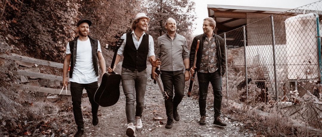 Vier Männer, jeder mit einer Gitarre, gehen gemeinsam auf einem Kiesweg. Sie tragen lässige Kleidung, einer trägt einen Hut und ein anderer eine Halskette. Dahinter ist ein Gebäude mit einem Zaun zu sehen.