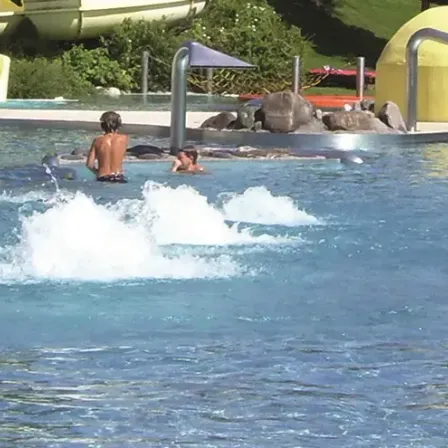 Mehrere Menschen schwimmen und planschen in einem Pool mit Wasserspiel. Ein Wasserrutsche ist im Hintergrund.