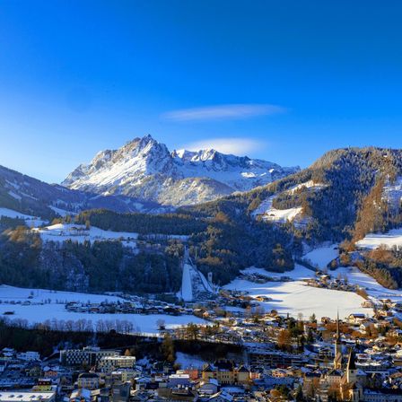 Luftaufnahme einer Stadt am Fuße eines Gebirgszuges mit blauem Himmel, schneebedeckten Gipfeln und klarem blauem Himmel.
