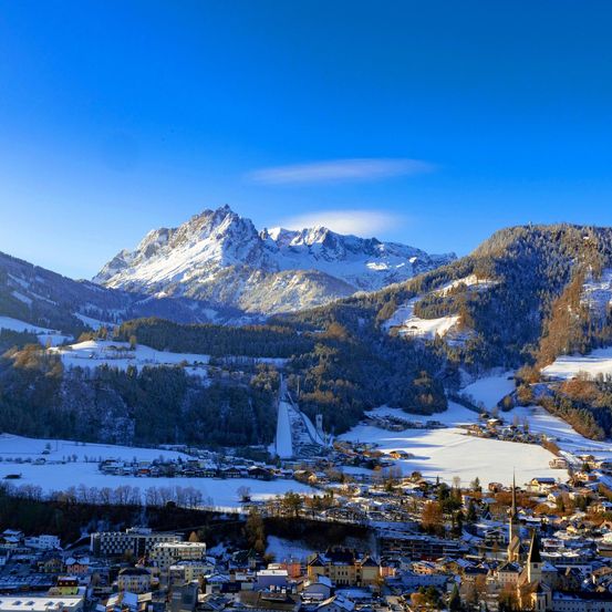 Luftaufnahme einer Stadt am Fuße eines Gebirgszuges mit blauem Himmel, schneebedeckten Gipfeln und klarem blauem Himmel.
