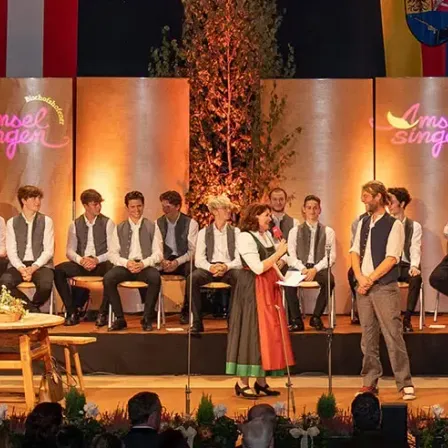Eine Gruppe von Menschen in traditionellen Kostümen steht auf einer Bühne mit einem Banner, das 'Insel Singer' sagt. Sie spielen Instrumente wie Harfe und Gitarre. Einige sitzen auf Stühlen, während andere stehen. Die Bühne ist mit Pflanzen und Blumen geschmückt.