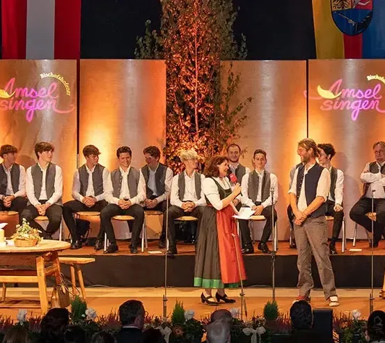 Eine Gruppe von Menschen in traditionellen Kostümen steht auf einer Bühne mit einem Banner, das 'Insel Singer' sagt. Sie spielen Instrumente wie Harfe und Gitarre. Einige sitzen auf Stühlen, während andere stehen. Die Bühne ist mit Pflanzen und Blumen geschmückt.