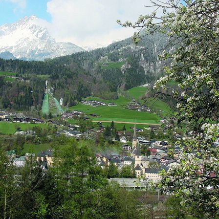Luftaufnahme einer Berglandschaft mit einer Stadt im Vordergrund. Die Stadt ist von üppigen grünen Feldern und dichten Wäldern umgeben. In der Ferne befindet sich eine Skisprungschanze, und der Himmel ist klar mit einigen Wolken.
