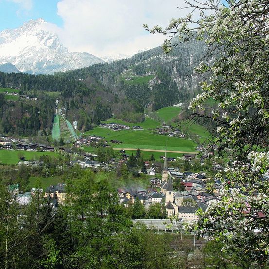 Luftaufnahme einer Berglandschaft mit einer Stadt im Vordergrund. Die Stadt ist von üppigen grünen Feldern und dichten Wäldern umgeben. In der Ferne befindet sich eine Skisprungschanze, und der Himmel ist klar mit einigen Wolken.