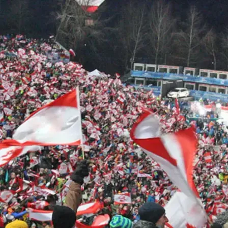 Eine große Menschenmenge schwenkt Flaggen bei einer Nachtveranstaltung. Einige Flaggen sind rot und weiß, andere blau und weiß. Die Menge ist rund um das Stadion versammelt, und einige Leute stehen auf den Geländern. In der Ferne befindet sich eine Bühne mit einem weißen Auto, das darauf geparkt ist.