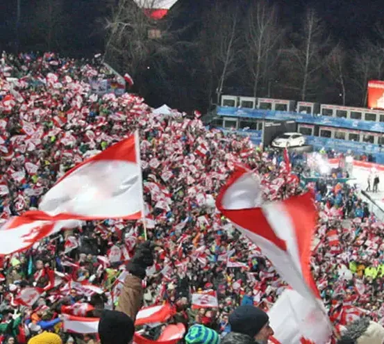 Eine große Menschenmenge schwenkt Flaggen bei einer Nachtveranstaltung. Einige Flaggen sind rot und weiß, andere blau und weiß. Die Menge ist rund um das Stadion versammelt, und einige Leute stehen auf den Geländern. In der Ferne befindet sich eine Bühne mit einem weißen Auto, das darauf geparkt ist.