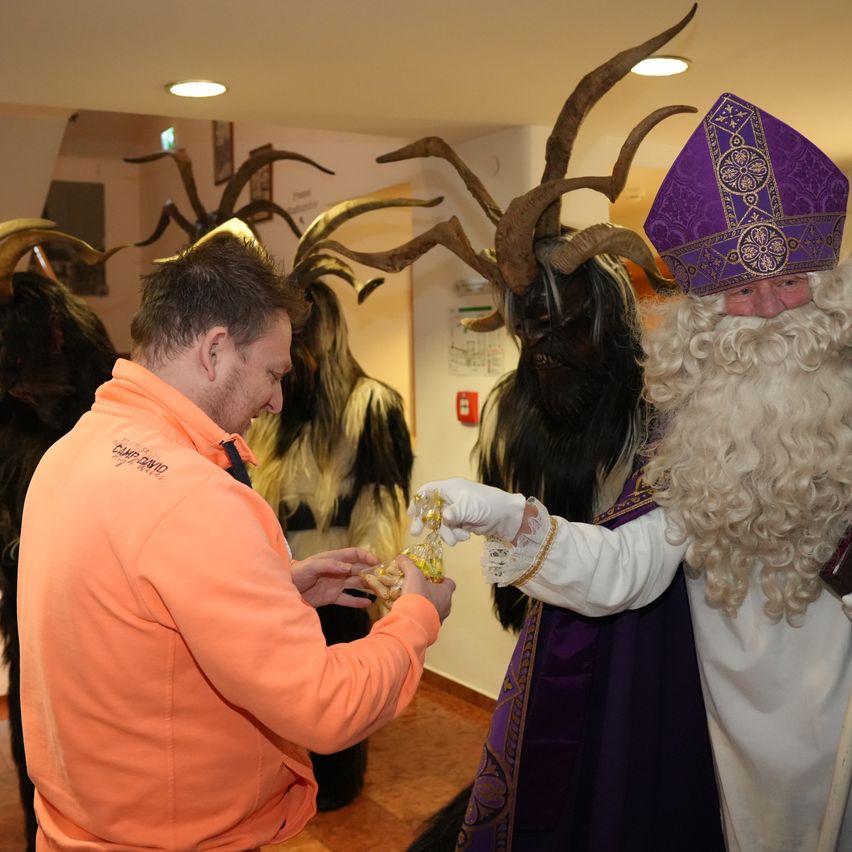 Ein Mann in einem orangefarbenen Sweatshirt erhält etwas von einer als Bischof gekleideten Person, mit einer Gruppe von Personen in Hörnermasken hinter ihnen.