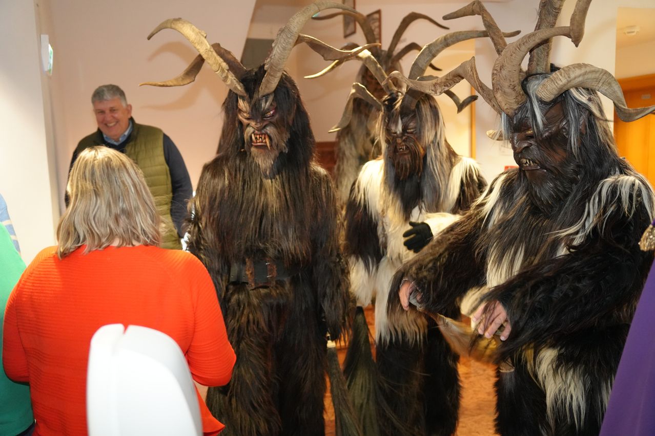 Vier Personen in haarigen Kostümen mit Hörnern und Masken stehen in einem Raum, einer hält eine große Hupe. Ein Mann und eine Frau beobachten sie von hinten.
