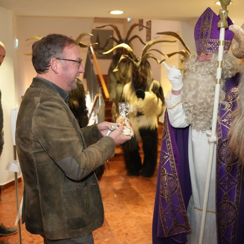 Ein Mann in einer braunen Jacke reicht einem als Weihnachtsmann verkleideten Menschen eine verpackte Leckerei. Dahinter stehen eine Gruppe von Menschen in Hörner-Kostümen in der Nähe einer Treppe.