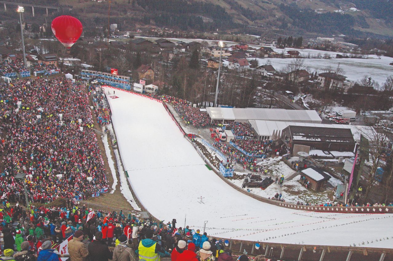 Ein Skisprungwettbewerb mit einer großen Zuschauermenge. Im Hintergrund schneebedeckte Hügel. Ein roter Heißluftballon ist sichtbar.
