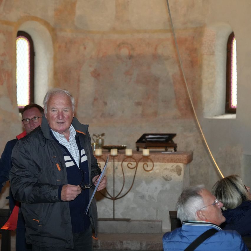 Ein älterer Mann steht in einer Kapelle, hält ein Papier in der Hand, mit zwei anderen, die hinter ihm sitzen. Die Kapelle hat einen Steinaltar mit Kerzen und einem Bogenfenster.