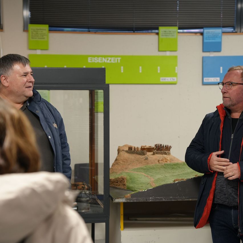 Zwei Männer stehen vor einer Vitrine mit einem Modell einer historischen Schlacht. Ein Mann trägt eine Brille und eine Jacke. Hinter ihnen befindet sich eine weiße Wand mit grünen und blauen Plakaten.