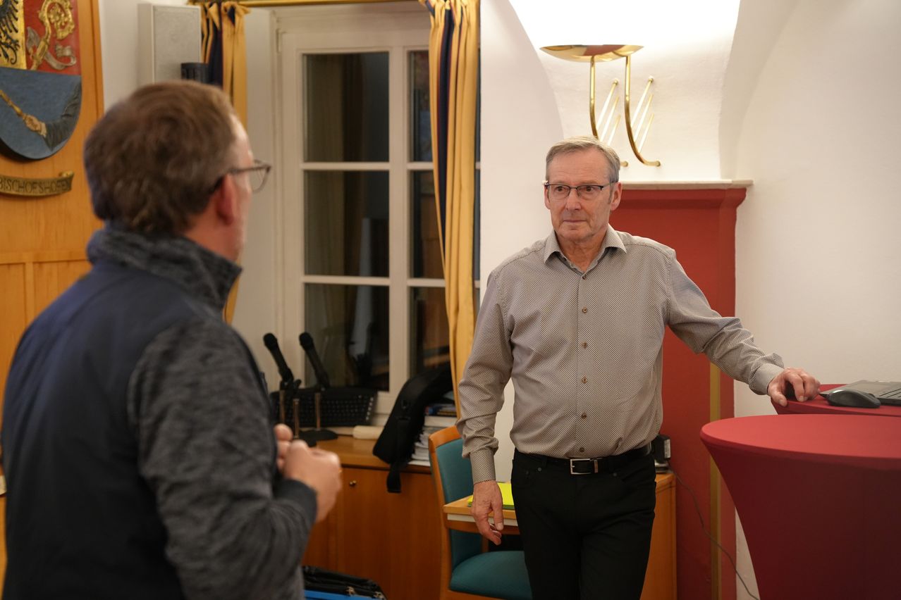 Zwei Männer sind in einem Raum, einer von ihnen schaut den anderen an, der zum Fenster schaut. Der Raum hat einen Schreibtisch mit Mikrofonen und Büchern.