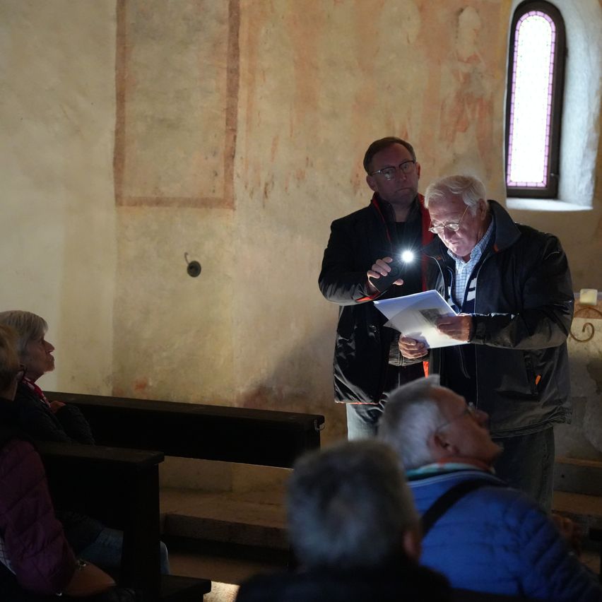 Zwei Männer stehen vor einer Menschenmenge in einem schwach beleuchteten Raum. Ein Mann hält eine Taschenlampe, während der andere ein Stück Papier liest. Der Raum hat Steinwände, ein Fenster und eine Holzbank.