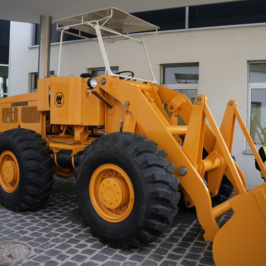 Ein gelber Liebherr-Bagger steht vor einem Gebäude. Das Fahrzeug hat eine große Frontschaufel und steht auf einer Kopfsteinpflasteroberfläche.