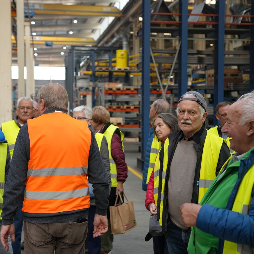 Eine Gruppe älterer Erwachsener in Warnwesten geht durch ein Lagerhaus, diskutiert und beobachtet die Umgebung.