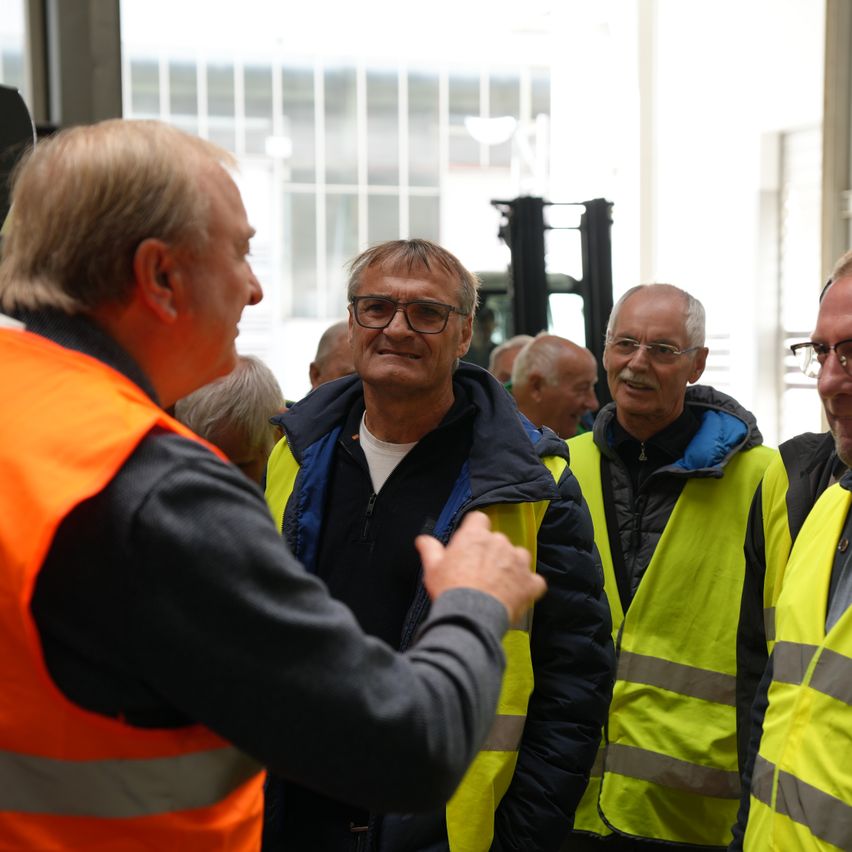 Eine Gruppe von Männern in Sicherheitswesten steht und unterhält sich in einer Fabrik oder einem Lager. Sie tragen Brillen und einige lächeln. Der Mann in der Mitte schaut den Mann auf der linken Seite an. Hinter ihnen steht ein Gabelstapler und eine Glaswand.