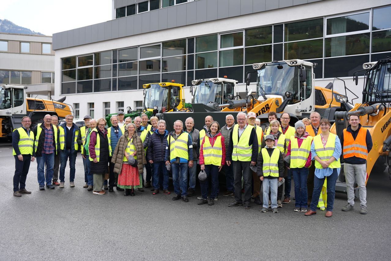 Eine Gruppe von Menschen in Warnwesten steht vor einem modernen Gebäude mit großen Fenstern. Hinter ihnen sind mehrere Baufahrzeuge geparkt.