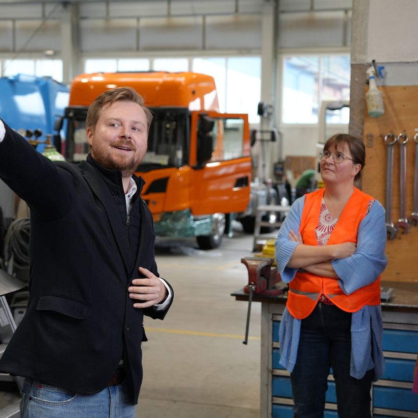 Ein Mann und eine Frau stehen in einer Werkstatt. Der Mann gestikuliert mit seinem Arm. Die Frau steht mit verschränkten Armen. Ein orangefarbener LKW steht hinter ihnen.