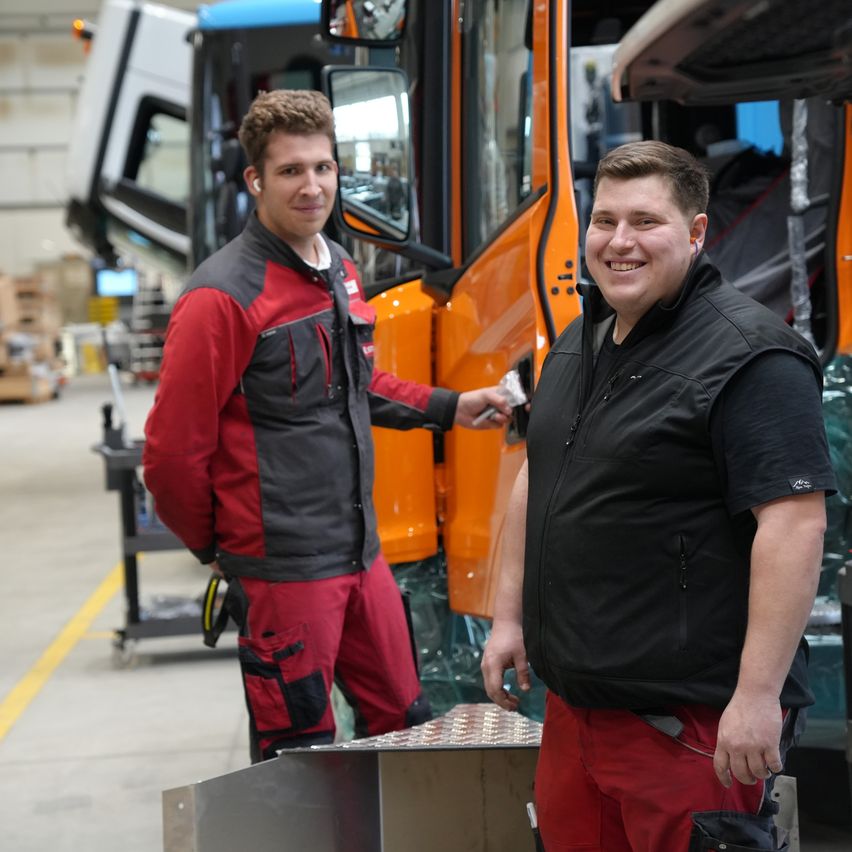 Zwei Männer stehen neben einem orangefarbenen Fahrzeug in einer Fabrik, einer trägt einen roten und schwarzen Overall, der andere eine schwarze Weste. Sie haben beide Werkzeuge in der Hand und scheinen zu lächeln.