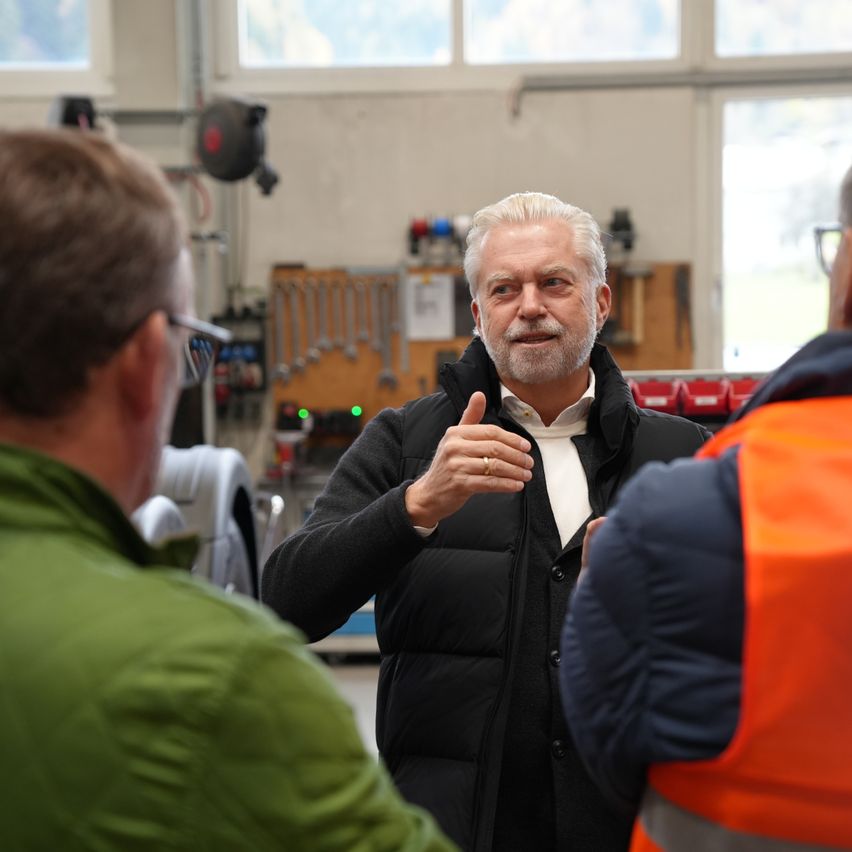 Ein älterer Mann mit weißem Haar und Bart steht in einer Werkstatt und spricht mit zwei Männern vor ihm. Werkzeuge und Geräte sind im Hintergrund zu sehen.