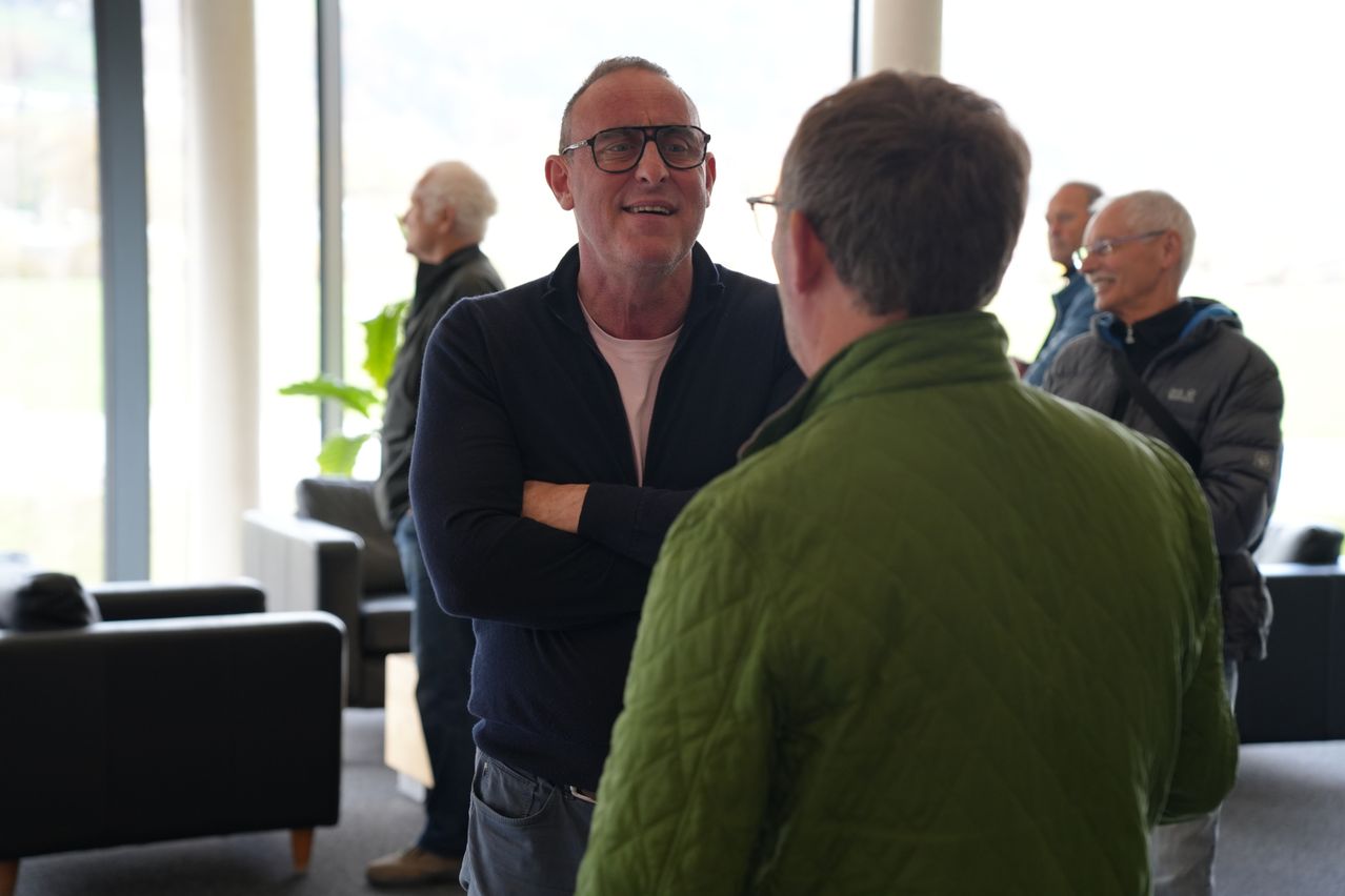 Ein erwachsener Mann mit Brille lächelt und spricht mit einem anderen Mann in einer grünen Jacke. Hinter ihnen steht eine Gruppe von Menschen in der Nähe eines großen Fensters.
