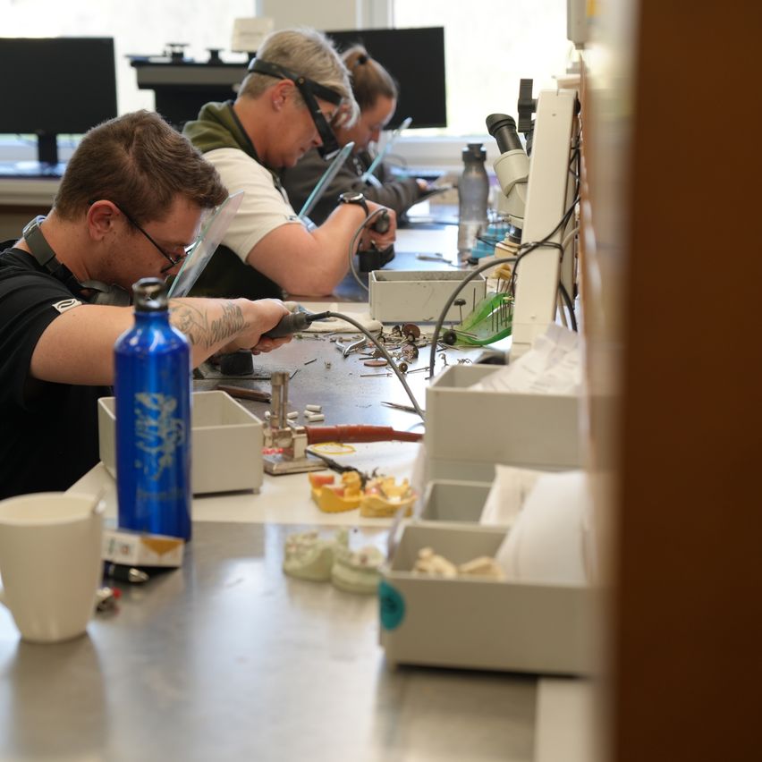 Drei Personen führen an einem Arbeitsplatz eine wissenschaftliche Aufgabe durch, unter Verwendung von Mikroskopen und Werkzeugen. Einer lötet, ein anderer beobachtet durch ein Mikroskop und der dritte arbeitet am Computer.