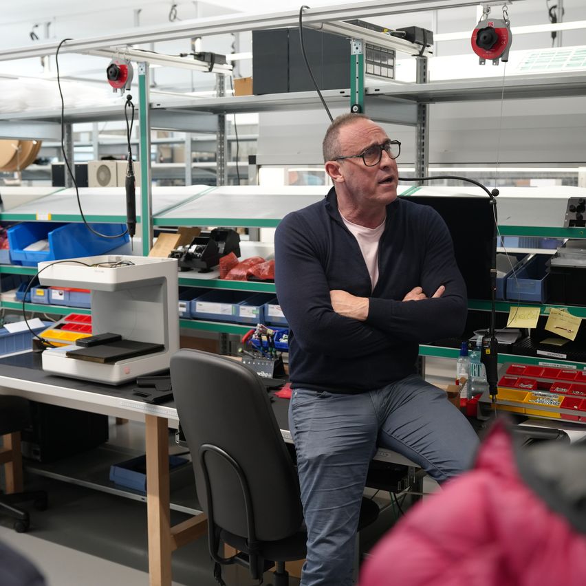 Ein Mann mit Brille steht in einer Fabrik mit verschränkten Armen. Hinter ihm befinden sich Regale mit Aufbewahrungsboxen und Ausrüstung. Eine Maschine mit schwarzem Sockel steht auf einem Schreibtisch.