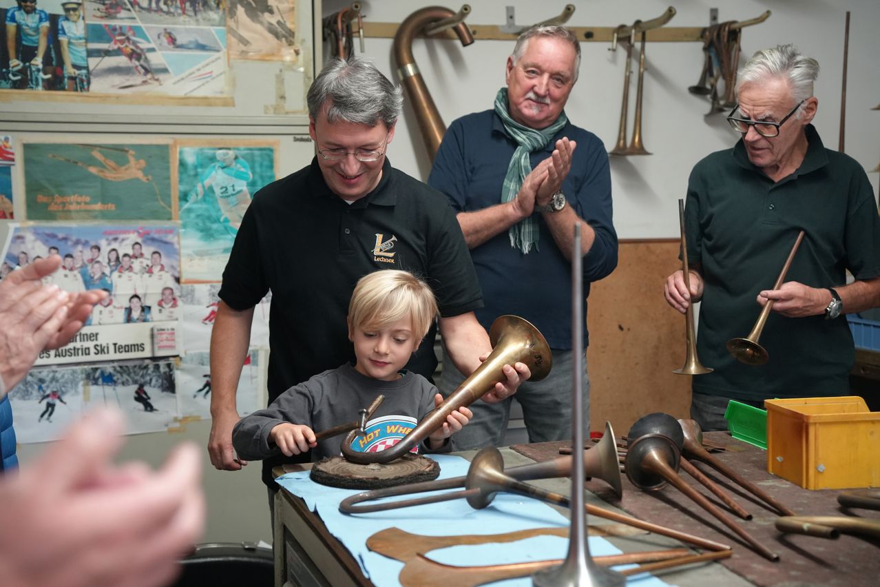 Ein kleiner Junge hält eine Trompete, während zwei Männer zusehen, einer lächelt und klatscht. Sie sind in einem Raum mit Musikinstrumenten an der Wand und auf einem Tisch.