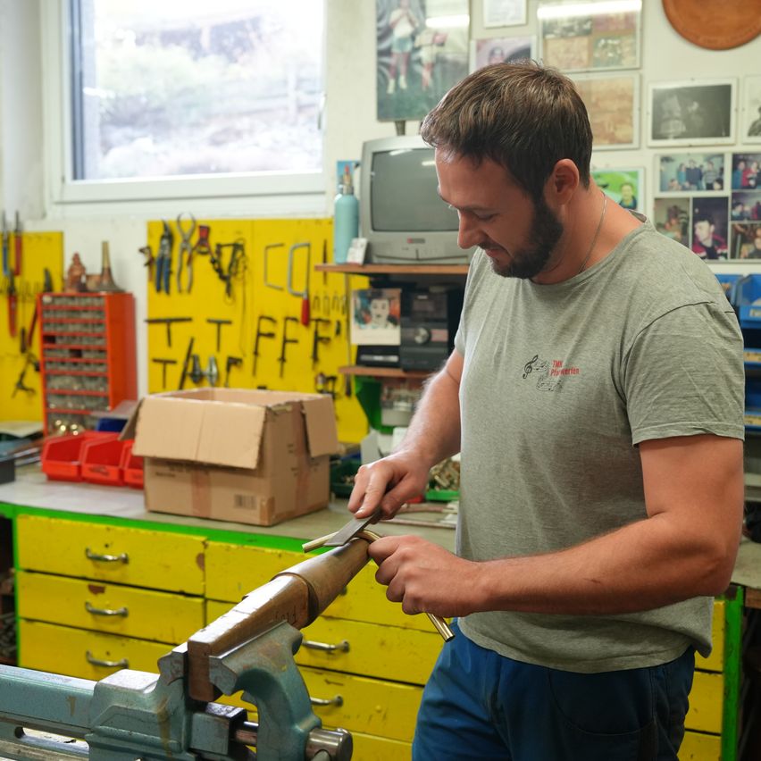 Ein Mann arbeitet in einer Werkstatt und benutzt ein Werkzeug auf einer Drehbank. Der Raum ist mit verschiedenen Werkzeugen, Lagerboxen und einem Fernseher auf einem Regal gefüllt. An der Wand hängen gerahmte Bilder.