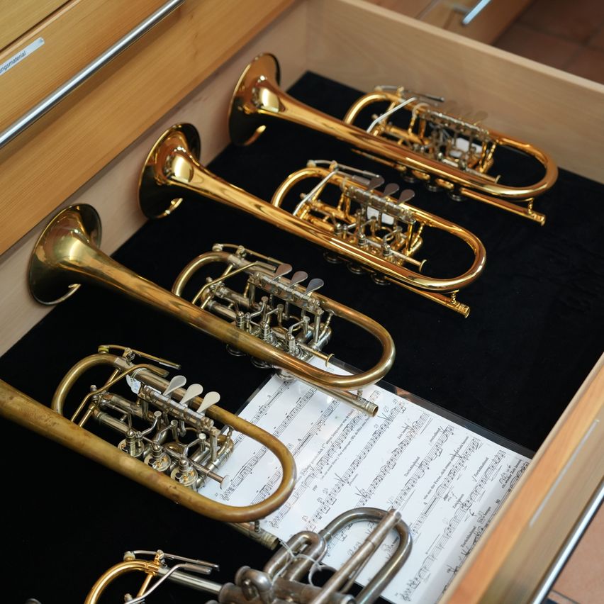 Ein hölzerner Schublade mit Messinginstrumenten. Vier Trompeten und ein Posaune sind ordentlich angeordnet. Die Schublade ist mit schwarzem Samt ausgekleidet. Musikalische Noten sind teilweise sichtbar.