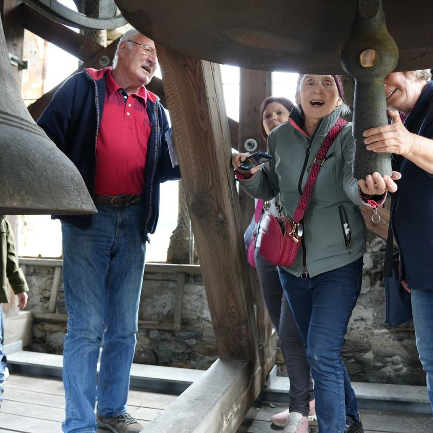 Eine Gruppe von Menschen steht in einem Glockenturm. Ein Mann lacht, während andere die Glocke berühren.