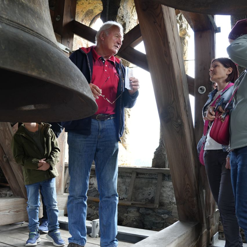 Ein älterer Mann in einem roten Shirt steht neben einer großen Glocke, hält ein Papier in der Hand und trägt eine Brille. Hinter ihm steht ein junger Junge in einer grünen Jacke und schaut zu. In der Nähe lächelt eine Frau in einer Jeansjacke und einer rosa Tasche zur Kamera.