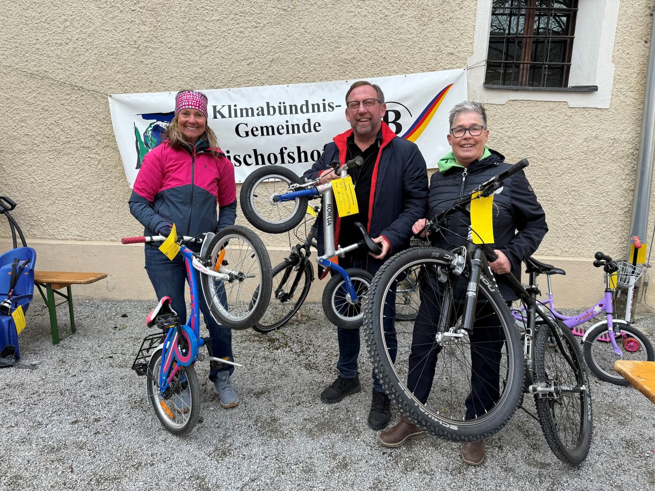 Drei Personen stehen neben Fahrrädern vor einem Gebäude. Ein Banner mit dem Text Klimabündnis-Gemeinschaft Bischofshofen hängt hinter ihnen. Sie lächeln und posieren für ein Foto.