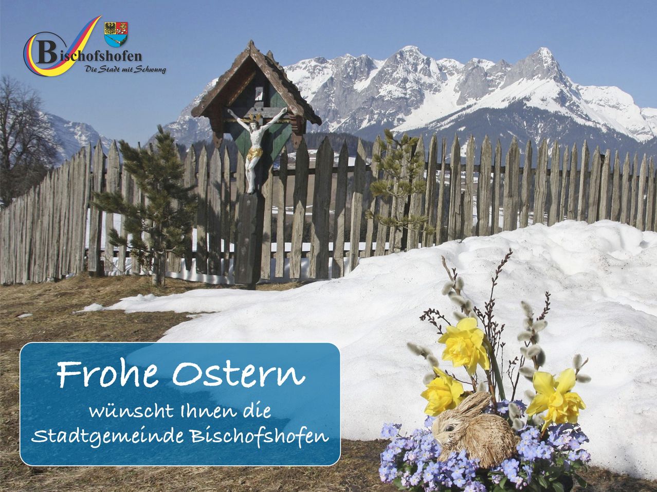 Ein Holzkreuz steht hinter einem Holzzaun, mit Schnee und Bergen im Hintergrund. Blumen und ein Hase sind im Vordergrund. Der Text sagt Frohe Ostern, wünscht Ihnen ein frohes Osterfest.