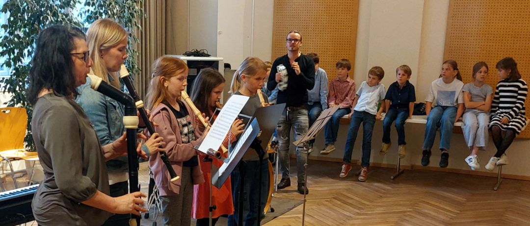 Eine Gruppe von Kindern spielt Musikinstrumente in einem Klassenzimmer, während ein Mann mit Brille hinter ihnen steht. Ein Kind hält eine Blockflöte, während andere Flöten spielen. Sie schauen auf Notenständer.